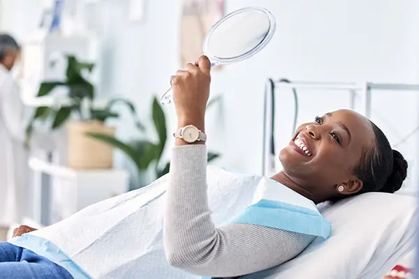 Smiling woman laying on dental chair, holding a mirror to her new dental implants at Mountain State Oral and Facial Surgery in Ashland, KY